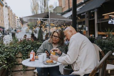 Male and female senior friends discussing over smart phone while having food at sidewalk cafe - MASF39767