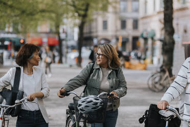 Senior female friends talking to each other while wheeling bicycles - MASF39786