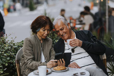 Senior woman sharing smart phone with elderly man holding coffee cup at sidewalk cafe - MASF39801