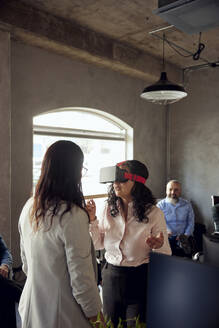 Geschäftsfrau, die einen VR-Simulator trägt, während sie von einer Kollegin im Büro gestikuliert wird - MASF39812