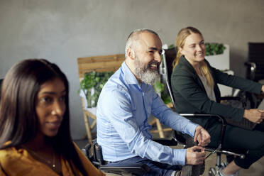 Happy bearded businessman with disability sitting amidst female colleagues in creative office - MASF39822