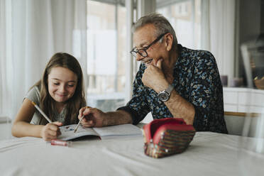 Lächelnder älterer Mann hilft seiner Enkelin bei den Hausaufgaben zu Hause - MASF39908