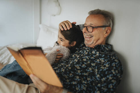 Cheerful senior man reading storybook with granddaughter on bed at home - MASF39928