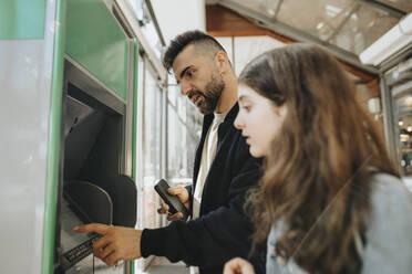 Vater benutzt einen Geldautomaten, während er mit seiner Tochter am Bahnhof steht - MASF39962