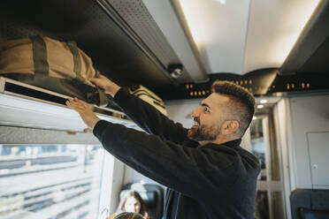 Seitenansicht eines lächelnden Mannes, der sein Gepäck in einem Zug auf ein Regal lädt - MASF39968