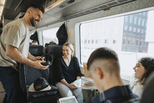 Lächelnder Mann nimmt Kopfhörer aus dem Koffer, während er im Zug mit seiner Familie spricht - MASF39970