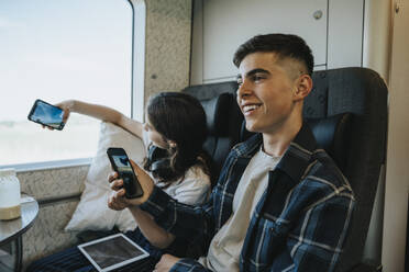 Glücklicher Junge, der sein Smartphone hält, während er auf einem Sitz im Zug sitzt - MASF39986
