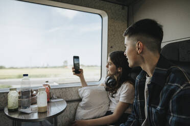 Junge schaut seine Schwester an, die im Zug mit dem Smartphone fotografiert - MASF39988