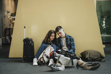 Boy and girl sharing smart phone while sitting with luggage against yellow wall - MASF40011