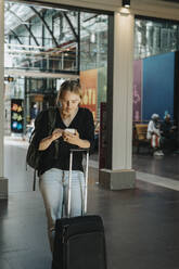 Frau benutzt Smartphone, während sie mit Gepäck am Bahnhof steht - MASF40022