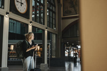 Nachdenkliche Frau mit Smartphone, die ihr Gepäck auf dem Bahnhof trägt - MASF40023