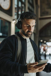 Man wearing bluetooth headphones holding smart phone at station - MASF40024