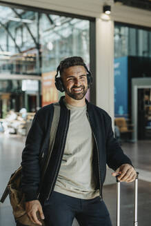 Porträt eines glücklichen Mannes mit Bluetooth-Kopfhörern, der mit seinem Gepäck am Bahnhof steht - MASF40025