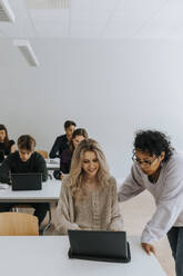 Teacher assisting young woman with laptop against students in classroom - MASF40044