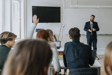 Männlicher Professor unterrichtet, während eine Studentin mit erhobener Hand im Klassenzimmer sitzt - MASF40053