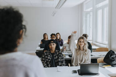 Junge Schülerin mit erhobener Hand, die bei Freunden sitzt und den Lehrer im Klassenzimmer anschaut - MASF40057