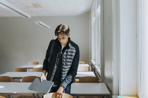 Teenage boy holding laptop while standing in classroom - MASF40095