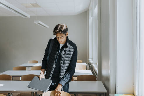 Teenage boy holding laptop while standing in classroom - MASF40095