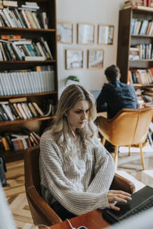 Blonde junge Frau, die einen Laptop benutzt, während sie in einer Bibliothek sitzt - MASF40101