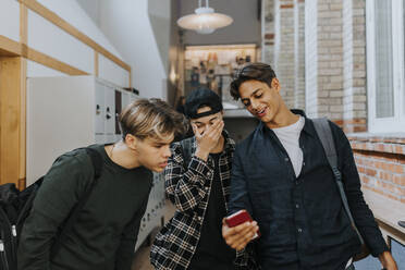 Teenager, der sein Smartphone mit Freunden teilt, während er durch den Schulkorridor geht - MASF40124
