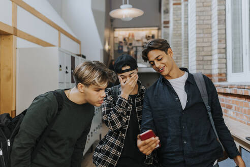 Teenage boy sharing smart phone with friends while walking in school corridor - MASF40124