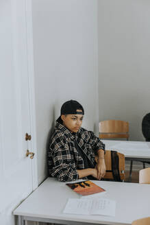 Nachdenklicher junger Mann sitzt am Schreibtisch und lehnt sich an eine weiße Wand im Klassenzimmer - MASF40139