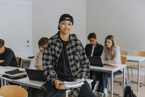 Porträt eines lächelnden jungen Mannes mit Buch am Schreibtisch im Klassenzimmer - MASF40150