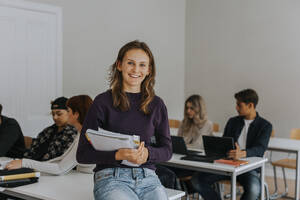 Porträt einer glücklichen jungen Studentin mit einem Buch, das auf einem Schreibtisch im Klassenzimmer liegt - MASF40155