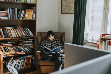 Junger Mann liest ein Buch, während er auf einem Sessel neben einem Bücherregal in einer Bibliothek sitzt - MASF40161