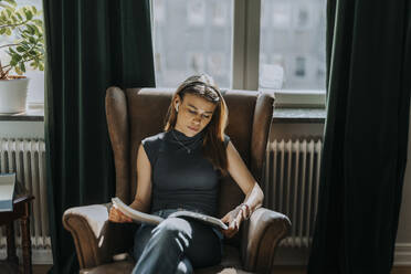 Teenage girl reading book while sitting on armchair against window at library - MASF40162