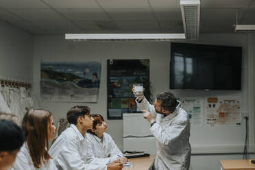 Männlicher Professor, der eine Chemikalie in einem Becherglas untersucht, während er einem Studenten im wissenschaftlichen Labor etwas erklärt - MASF40166