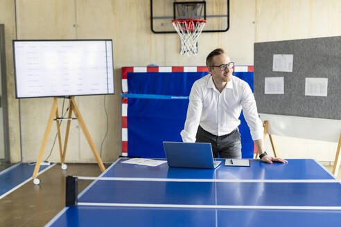 Glücklicher Geschäftsmann mit Laptop, der sich auf einen Tennistisch in einem modernen Büro stützt - PESF04040