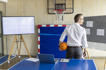 Ein lächelnder Geschäftsmann steht mit einem Basketball vor einem Basketballkorb und einem Fernsehbildschirm - PESF04042