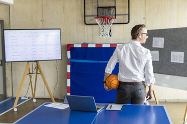 Smiling businessman standing with basketball in front of hoop and television screen - PESF04042