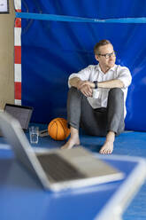 Smiling businessman sitting with coffee cup near basketball and laptop in office - PESF04047