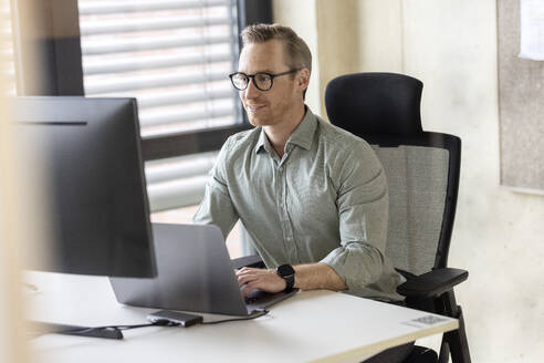 Lächelnder Geschäftsmann bei der Arbeit an Laptop und Desktop-PC im Büro - PESF04083