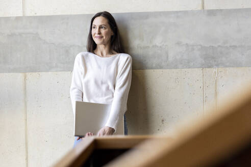 Thoughtful mature businesswoman standing with laptop in front of wall - PESF04108