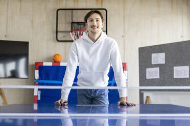 Smiling businessman leaning on tennis table in office - PESF04114