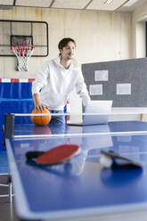 Smiling young businessman standing with basketball and laptop at tennis table - PESF04115