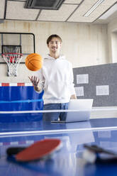 Smiling businessman standing with basketball and laptop at tennis table - PESF04116