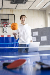 Lächelnder Geschäftsmann stehend mit Basketball und Laptop am Tennistisch - PESF04116