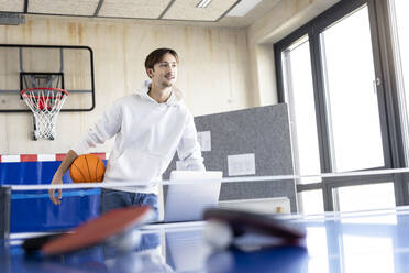 Smiling young trainee standing with basketball in office - PESF04118