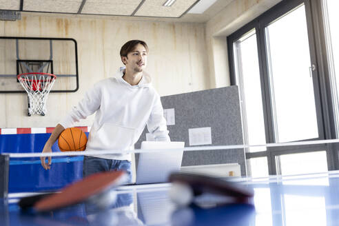 Lächelnder junger Auszubildender mit Basketball im Büro stehend - PESF04118