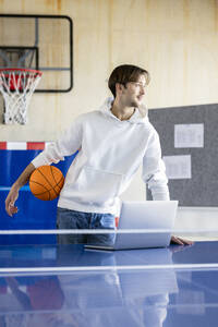 Junger Auszubildender mit Basketball und Laptop im Büro stehend - PESF04119