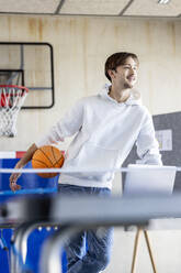 Happy young trainee standing with basketball and laptop in office - PESF04120