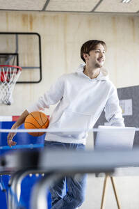 Glücklicher junger Auszubildender mit Basketball und Laptop im Büro stehend - PESF04120