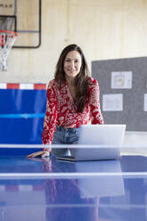 Happy mature businesswoman leaning with laptop on tennis table - PESF04124