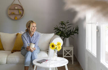 Smiling woman with tea cup on sofa looking thorough window at home - SVKF01655