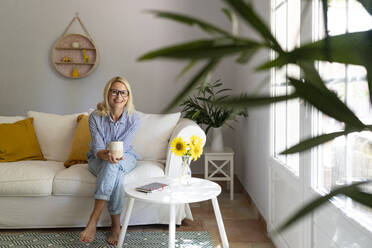 Smiling mature woman with tea cup sitting on sofa in living room at home - SVKF01657