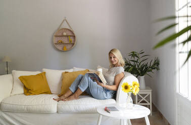 Mature woman with book having tea on sofa at home - SVKF01661